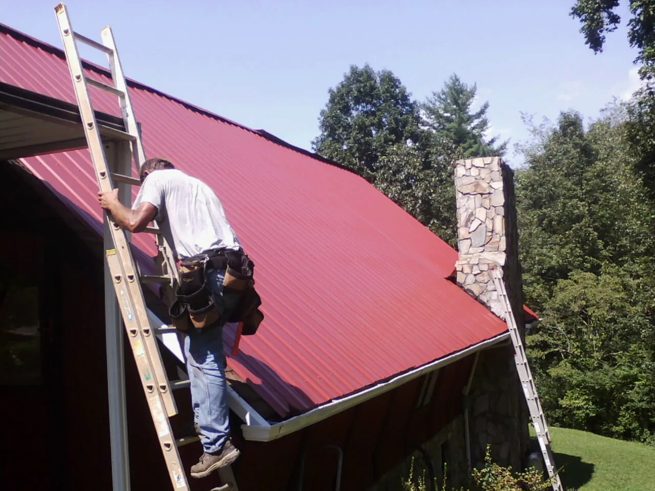 Roofer on a ladder beside a red metal roof and stone chimney — Soffit & Fascia Repair by Roofing Brothers in Scottsbluff